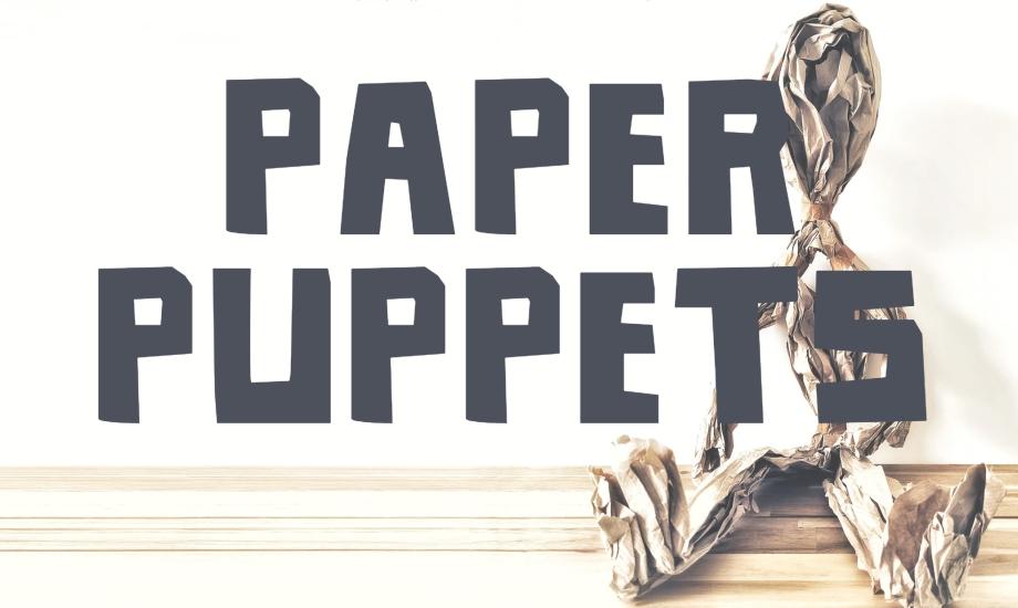 A paper puppet sat against a wall with a paper puppet writing across the photo